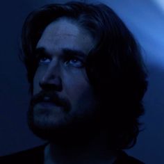 a man with long hair and blue eyes stares into the distance in a dark room