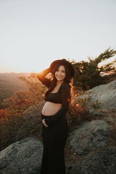 a pregnant woman standing on top of a rock in front of the sun wearing a hat