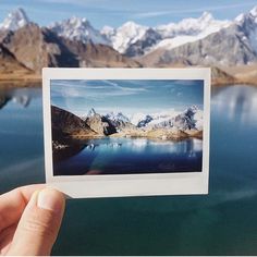 a person holding up a polaroid photo in front of some mountains and blue water