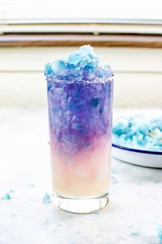 a glass filled with blue and purple liquid next to a bowl full of ice cubes