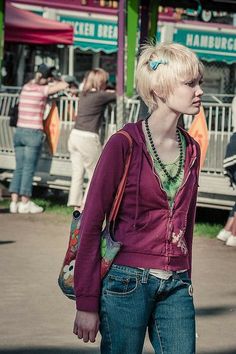 a woman with blonde hair is standing in front of a fairground and looking off into the distance