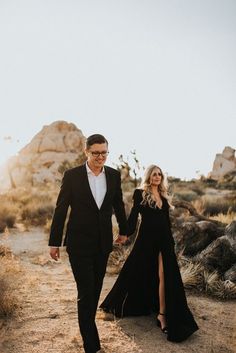 a man and woman walking down a dirt road holding each other's hand as they walk towards the camera