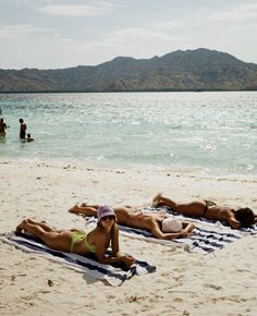 two women in bikinis laying on towels at the beach with mountains in the background