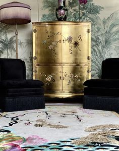 two black chairs and a gold cabinet in a room with floral wallpaper on the walls