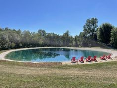 there are red chairs around the pool in the field by the water and trees behind it