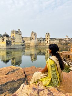 a woman sitting on top of a rock next to a body of water with buildings in the background