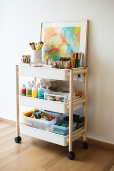 a painting and art supplies stand on wheels in front of a white wall with wooden flooring