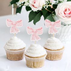 three cupcakes with pink bows on them are sitting next to a bouquet of flowers