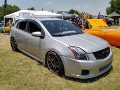 a silver car parked in the grass near other cars and people at an outdoor event