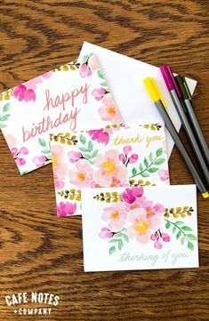 three cards with flowers on them sitting next to some paintbrushes and paper pens