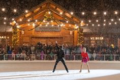 two people standing in front of a building at night with lights on the roof and snow falling all around them