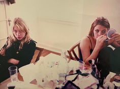 two women sitting at a table with plates and cups in front of them, one holding a cup