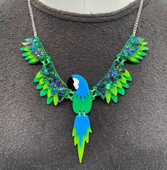 Polly Parrot 🦜 necklace is a fun quirky piece. It's a statement piece that will look great with any outfit.  The necklace is delicately made with green, blue and our fabulous glitter green acrylic. The wings are linked together using jump rings and connected to a curb chain.  If you have any questions drop us a line!💌 We are more than happy to help! 😁 To clean your necklace you can simply wipe with a glasses cloth or soft cloth to remove finger marks. Acrylic is a fragile material so please store in a jewellery box to prevent damage.  As ever for safety please keep away from children. Handmade Green Novelty Necklace, Trendy Resin Necklaces For Party, Green Novelty Resin Jewelry, Unique Green Resin Necklaces, Unique Green Resin Necklace, Quirky Handmade Green Jewelry, Novelty Green Necklace For Gifts, Green Novelty Necklace For Gift, Unique Green Necklace For Gift