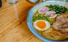 a bowl of ramen with meat, eggs and greens on the side sitting on a wooden table