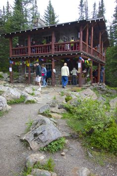 several people standing in front of a wooden cabin on top of a rocky hill near trees