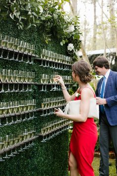 a man and woman standing next to a wall covered in cups with wine glasses on them