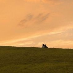 two people are sitting in the grass on top of a hill with an orange sky behind them