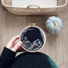 a person is holding up a small embroidery project in front of a basket and pumpkins