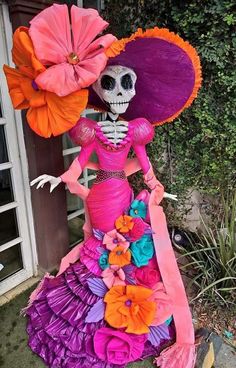 a skeleton wearing a colorful dress and hat with flowers on it's head is standing in front of a house