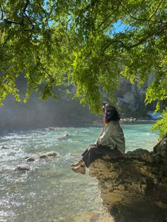 a man sitting on top of a rock next to a river under a green leafy tree