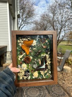 a person holding up a framed painting with butterflies and pine cones on the ground in front of a house