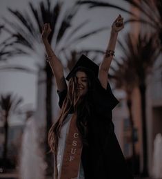 a woman with her arms in the air wearing a graduation cap and gown while standing next to a fountain