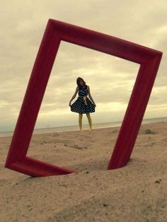 a woman standing on top of a sandy beach next to an empty red frame in the sand