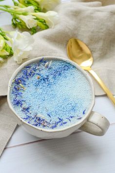 a white cup filled with blue liquid next to a gold spoon and flowers on a table