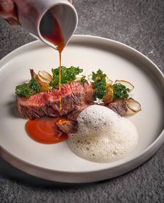 someone is pouring sauce on a steak with vegetables and potatoes in a white plate that sits on a gray table