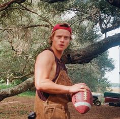 a man in overalls holding a football under a tree