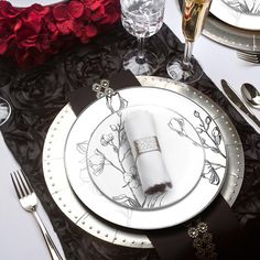 a place setting with black and white plates, silverware and red flowers on the table