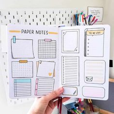 a person holding up a planner book in front of a desk with pens and pencils