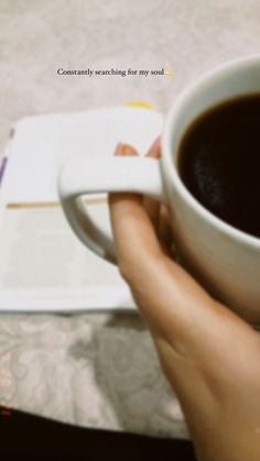 a person is holding a cup of coffee in their left hand and an open book on the table