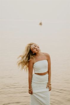 a woman in a white dress is standing by the water with her hair blowing in the wind