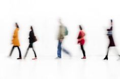 blurry image of people walking in a line on a white floor with one person wearing an orange jacket