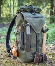 a green backpack with tools attached to it in the grass and trees behind it is an assortment of utensils