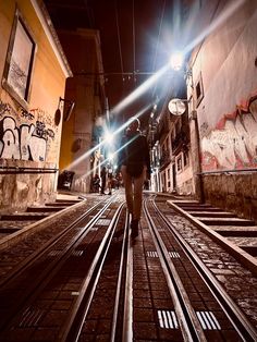 a man walking down a street at night with graffiti on the walls and tracks in front of him