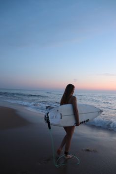 a woman holding a surfboard on top of a beach next to the ocean at sunset