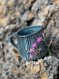 a blue cup sitting on top of a rock next to some pink flowers and leaves