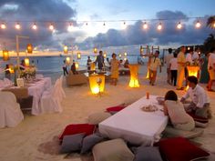 people sitting on the beach at night with lanterns strung over them and tables set up for dinner