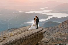 a bride and groom standing on top of a mountain with the sun setting in the background