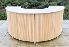 a curved wooden table sitting on top of a tile floor next to a green bush