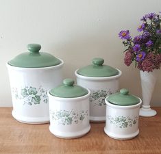 three white and green canisters with flowers in the vase next to them on a wooden table