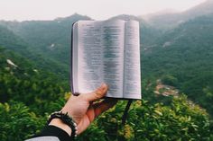 a person is holding an open book in their hand with mountains in the back ground