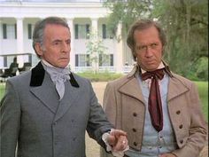 two men standing next to each other in front of a white house