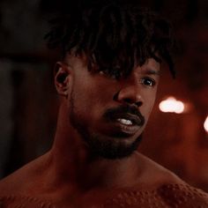 a shirtless man with dreadlocks stares at the camera in a dimly lit room