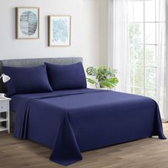 a bed with blue sheets and pillows in a white room next to a window on a wooden floor