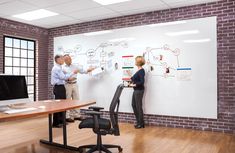 two people standing in front of a whiteboard with diagrams on it and another person sitting at a desk