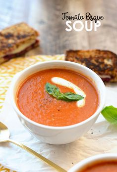 a bowl of tomato basil soup with grilled cheese on the side and toasted bread in the background