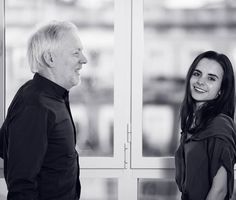 an older man and woman standing next to each other in front of large glass windows